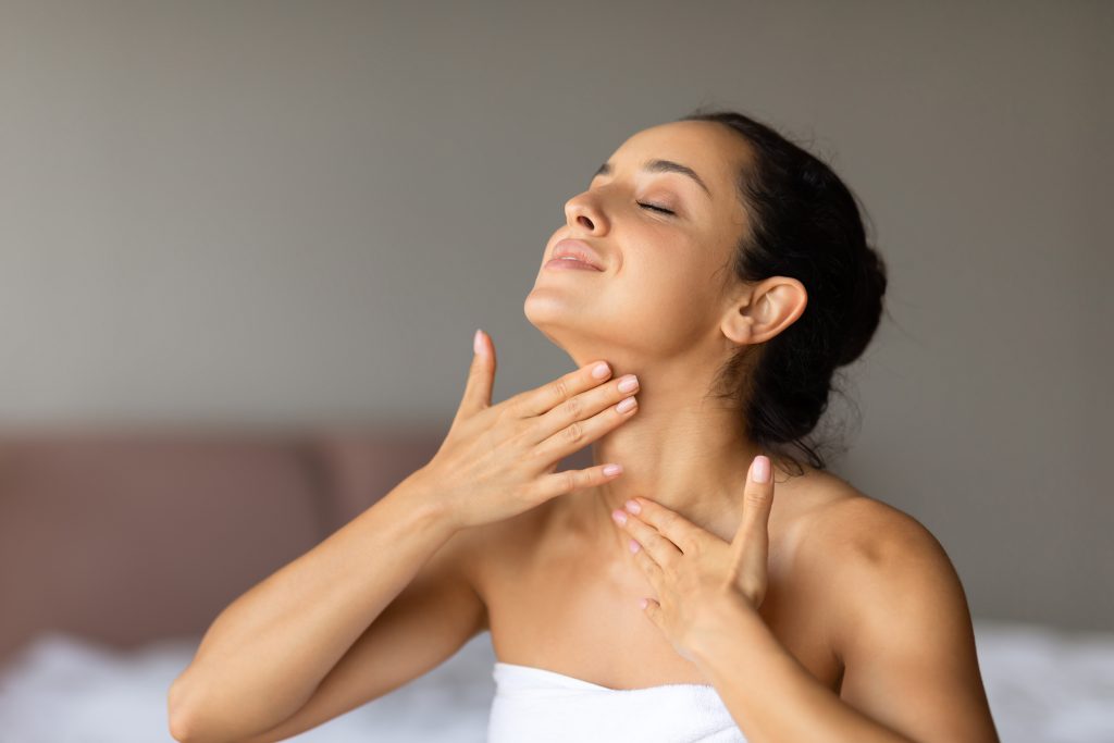 Happy caucasian lady applies moisturizer massaging and touching her neck, promoting skincare and self pampering, sitting wrapped in white soft towel at modern bedroom indoors