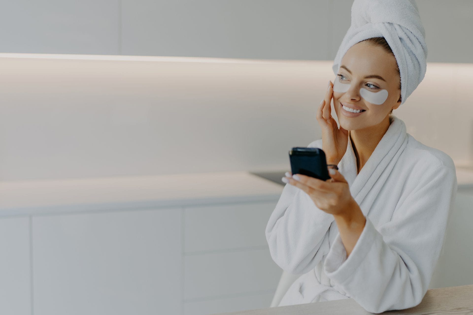 Happy smiling European woman applies moisturising patches under eyes touches face gently enjoys skin care routine dressed in white bathrobe looks somewhere aside poses at kitchen. Beauty concept Happy smiling European woman applies moisturising patches under eyes touches face gently enjoys skin care routine dressed in white bathrobe looks somewhere aside poses at kitchen. Beauty concept