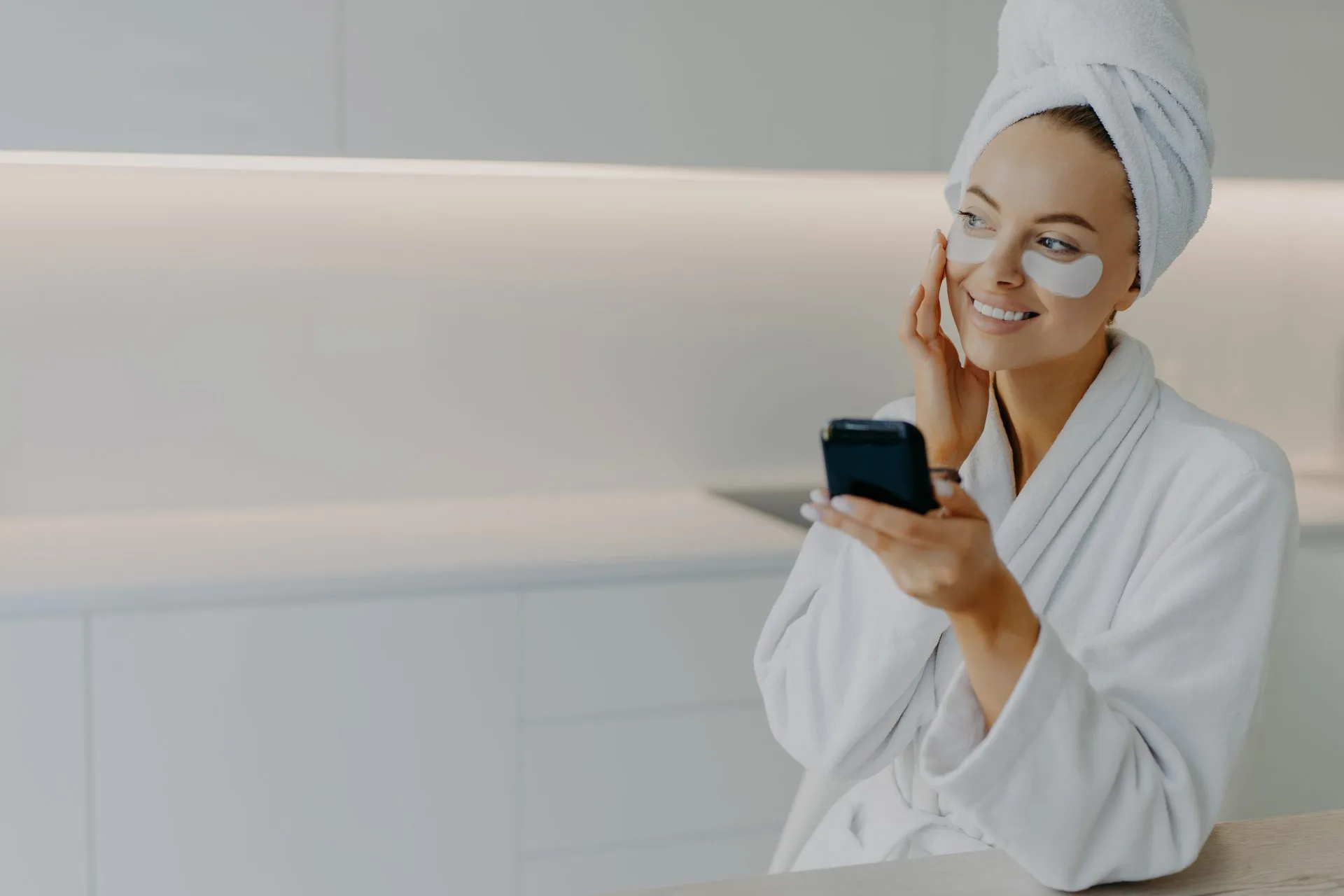 Happy smiling European woman applies moisturising patches under eyes touches face gently enjoys skin care routine dressed in white bathrobe looks somewhere aside poses at kitchen. Beauty concept